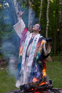 Rev. Jan, dressed in multicolored robe, stands in front of fire with one hand raised pointing to the sky.