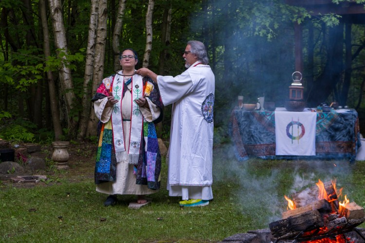 Rev. Drum (dressed in white robes) holds sickle to throat of Rev. Jan (dressed in multi-colored robes)